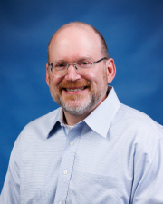 A man with glasses smiling for a photo with a blue background behind him. 